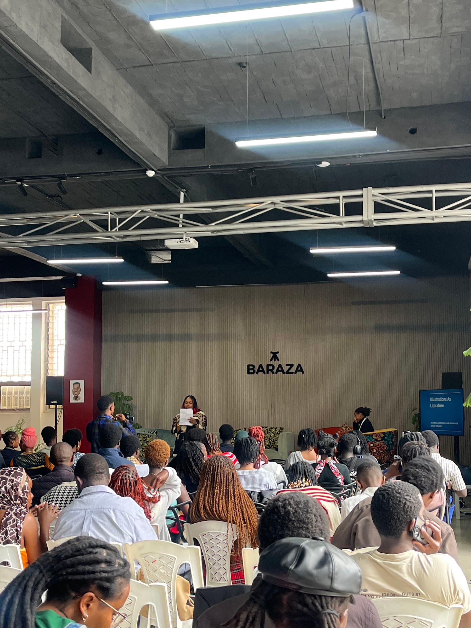 A seated audience in the main hall facing the stage beneath the Baraza sign
